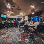 Ein Mann spielt Poker an einem Tisch in einem Casino. Er hat eine ernste Miene und hält Karten in der Hand, während er von anderen Spielern umgeben ist. Die Umgebung ist elegant und es herrscht eine
