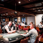 Ein Mann spielt Poker an einem Tisch mit grünem Filz. Er hält Karten in der Hand und hat einen ernsten Blick. Um ihn herum sitzen andere Spieler, die auf ihre Karten schauen. Das Bild