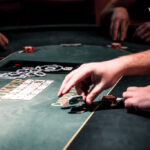 Ein professioneller Pokertisch mit grünem Filz, umgeben von Spielern, die Karten halten und Pokerchips stapeln. Ein Spieler mit Sonnenbrille und Kappe betrachtet konzentriert seine Hand,