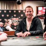 Ein Poker-Tisch mit grüner Filzdecke, umgeben von Spielkarten, Pokerchips und Getränken. Drei Personen sitzen um den Tisch herum und konzentrieren sich auf ihre Karten
