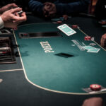 Ein Mann sitzt an einem Pokertisch mit grüner Filzdecke und hält eine Hand mit Spielkarten. Er trägt ein dunkles Hemd und hat eine ernste Miene. Im Hintergrund sind einige Chips