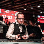 Ein Mann in einem dunklen Anzug und einer roten Krawatte sitzt an einem Pokertisch und hält Karten in der Hand. Er hat eine ernste Miene und konzentriert sich auf das Spiel. Im H