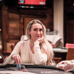 Ein Mann in einem schwarzen Anzug und einer roten Krawatte sitzt an einem Pokertisch und hält seine Karten in der Hand. In der Mitte des Tisches liegen Pokerchips und Spielkarten. Im