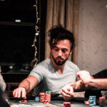Ein Mann sitzt an einem Pokertisch und hält seine Karten in der Hand, während er konzentriert und ernsthaft auf das Spielgeschehen schaut. Um ihn herum sind Pokerchips und Spielkarten
