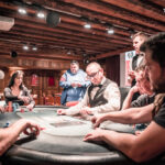 Ein Mann in einem schwarzen Anzug sitzt an einem Pokertisch und hält mit ernstem Gesichtsausdruck seine Karten. Im Hintergrund sind andere Spieler und Pokerchips zu sehen.