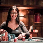 Ein Mann und eine Frau sitzen an einem Pokertisch, konzentriert auf ihre Karten schauend. Die Frau hat eine Hand am Kinn, während der Mann mit den Fingern auf den Chips vor sich tippt.
