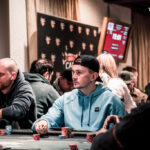 Ein Mann in einem dunklen Anzug und einer roten Krawatte sitzt an einem Pokertisch und hält seine Karten in der Hand, während er konzentriert auf den Tisch schaut. Um ihn herum sit