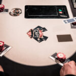 Ein Mann in einem dunkelblauen Anzug und einer weißen Hemdbluse sitzt an einem Pokertisch und hält in seiner Hand Pokerkarten. Er schaut konzentriert auf seine Karten, während im Hinter