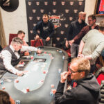 Ein professioneller Pokertisch mit grünen Filz, Karten und Chips, umgeben von vier Personen, die konzentriert Poker spielen.