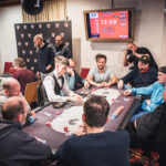 Ein Mann spielt Poker mit Freunden an einem Tisch, während er mit einem ernsten Gesichtsausdruck seine Karten betrachtet. Im Hintergrund sind weitere Spieler und Pokerchips auf dem Tisch zu sehen.