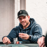 Ein Mann sitzt an einem Pokertisch und hält zwei Spielkarten in der Hand, während er konzentriert auf seine Gegner schaut. Hinter ihm sind Pokerchips und Spielkarten auf dem Tisch verteilt