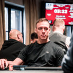 Ein Mann spielt Poker an einem Tisch mit grünem Filz und hält Karten in der Hand, während er konzentriert auf die Mitte des Tisches schaut. Im Hintergrund sind Chips und Geträn