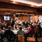 Ein Mann in einem schwarzen Anzug sitzt an einem Pokertisch, umgeben von anderen Spielern. Er hält Pokerkarten in der Hand und schaut konzentriert auf den Tisch. Die Atmosphäre ist span
