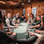 Ein Tisch mit einem grünen Pokertuch, umgeben von Spielkarten, Pokerchips und Getränken. Mehrere Personen sitzen um den Tisch und spielen Poker.