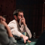 Ein Mann in einem schwarzen Anzug und einer schwarzen Sonnenbrille spielt Poker an einem Tisch in einem eleganten Casino. Er hält Karten in der Hand und schaut konzentriert auf den Tisch. Um