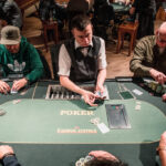 Ein professioneller Pokertisch mit grünem Filz, umgeben von Stühlen und Chips. Ein Stapel Spielkarten und ein Glas Whisky auf dem Tisch. Im Hintergrund sind unscharfe Spieler zu sehen.