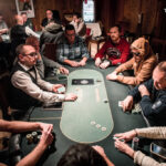 Ein Tisch mit vier Personen, die Poker spielen. Der Tisch ist mit Pokerchips und Spielkarten bedeckt, während die Spieler ernsthaft ihre Karten betrachten.