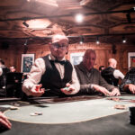 Ein Mann und eine Frau sitzen an einem Pokertisch und spielen eine Partie Texas Hold'em. Der Mann hat eine Hand mit Pokerchips vor sich und schaut konzentriert auf seine Karten, während die Frau ihn aufmer