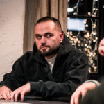 Ein Mann sitzt an einem Pokertisch, hält Spielkarten in der Hand und betrachtet sie konzentriert. Um ihn herum sind Pokerchips und andere Spieler zu sehen.