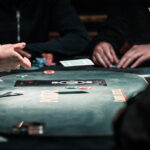 Ein Mann sitzt an einem Pokertisch und hält zwei Spielkarten in der Hand, während er konzentriert auf seine Gegner schaut. Eine Stapel von Pokerchips liegt vor ihm auf dem Tisch.