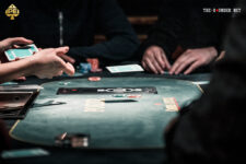 Ein Mann sitzt an einem Pokertisch und hält zwei Spielkarten in der Hand, während er konzentriert auf seine Gegner schaut. Eine Stapel von Pokerchips liegt vor ihm auf dem Tisch.