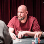 Ein Pokertisch mit grünem Filz, umgeben von Spielern, die Karten halten und Pokerchips stapeln. Ein Spieler hat gerade gewonnen und jubelt, während die anderen ihre Karten betrachten.