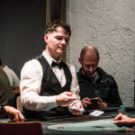 Ein Mann sitzt an einem Pokertisch, hält Karten in der Hand und schaut konzentriert auf seine Gegner. Die Szene spielt in einem eleganten Raum mit gedämpftem Licht und dezentem