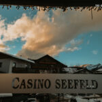 Ein Mann sitzt an einem Pokertisch und hält seine Karten in der Hand, während er konzentriert auf das Spiel schaut. Andere Spieler sind im Hintergrund zu sehen.