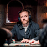 Ein Mann spielt Poker an einem Tisch mit grüner Filzdecke. Er hält Karten in der Hand und schaut konzentriert auf seine Chips. Im Hintergrund sind weitere Spieler und Gläser mit Get