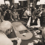 Ein Mann und eine Frau sitzen an einem Pokertisch und spielen konzentriert eine Runde Poker. Der Mann hat eine Hand mit Chips vor sich liegen, während die Frau Karten hält. Im Hintergrund sind weitere Spieler
