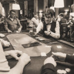 Ein Mann sitzt an einem Pokertisch und hält zwei Karten in der Hand, während er konzentriert auf die Gemeinschaftskarten schaut. Um ihn herum sind Chips und Getränke zu sehen.