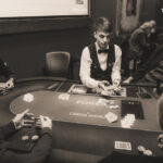Ein Mann spielt Poker an einem Tisch mit grüner Filzdecke. Er hält Karten in der Hand und betrachtet konzentriert seine Chips. Im Hintergrund sind weitere Spieler und ein Stapel Pokerch