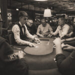 Ein Mann sitzt an einem Pokertisch und hält Karten in der Hand. Er schaut konzentriert auf seine Karten, während andere Spieler im Hintergrund zu sehen sind. Der Tisch ist mit Chips und Getr