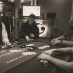 Ein Mann und eine Frau sitzen an einem Pokertisch und halten ihre Karten. Sie schauen konzentriert auf den Tisch. Im Hintergrund sind Pokerchips und Getränke zu sehen.