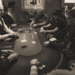 Ein Mann sitzt an einem Pokertisch mit grüner Filzdecke, umgeben von Spielkarten, Jetons und einem Stapel Pokerchips. Er hält zwei Karten in der Hand und schaut konzentri
