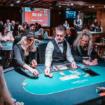 Ein Mann sitzt an einem Pokertisch und hält zwei Spielkarten in der Hand. Die Karten sind nicht sichtbar, aber er scheint konzentriert und fokussiert zu sein. Im Hintergrund sind