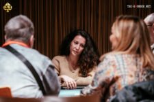 Ein Mann in einem eleganten Anzug sitzt an einem Pokertisch und hält seine Hand mit Spielkarten. Im Hintergrund sind weitere Spieler und Spielchips zu sehen.