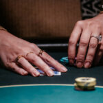 Ein Mann spielt Poker an einem Tisch in einem eleganten Casino. Er hält Karten in der Hand und betrachtet konzentriert seine Chips. Im Hintergrund sind andere Spieler und ein Croupier zu sehen.