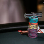 Ein Mann und eine Frau sitzen an einem Pokertisch und spielen ein Kartenspiel. Der Mann hat einen Stapel Chips vor sich, während die Frau ihre Karten betrachtet. Im Hintergrund sind weitere Spieler und eine Casino-U