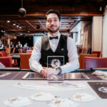 Ein Mann sitzt an einem Pokertisch und hält eine Hand mit Spielkarten. Er hat einen konzentrierten Gesichtsausdruck und trägt eine schwarze Kappe. Im Hintergrund sind weitere Pokerspi