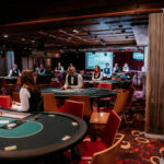 Ein Mann sitzt an einem Pokertisch und hält zwei Spielkarten in der Hand. Er schaut konzentriert auf den Tisch, umgeben von Pokerchips und anderen Spielern im Hintergrund.