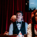 Ein Mann mit Sonnenbrille und Hut sitzt an einem Pokertisch und hält seine Karten in der Hand, während er konzentriert auf seine Gegner blickt. Die Chips stapeln sich vor ihm auf dem T