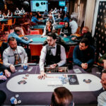 Ein professioneller Pokertisch mit grünem Filz, umgeben von Spielern, die Karten halten und Pokerchips stapeln. Ein Spieler in der Mitte des Bildes hat gerade gewonnen und jubelt darüber.