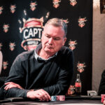 Ein Mann in einem eleganten Anzug und einer Fliege sitzt an einem Pokertisch und hält zwei Karten in der Hand. Er schaut konzentriert auf seine Karten, während im Hintergrund andere Spieler und