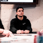 Ein Mann in einem dunkelblauen Anzug sitzt an einem Pokertisch und hält eine Hand mit Spielkarten. Er schaut konzentriert auf seine Karten, während die anderen Spieler im Hintergrund verschwommen