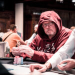Ein Mann mit einem roten Hemd und schwarzen Kopfhörern sitzt an einem Pokertisch und hält eine Hand mit Spielkarten. Im Hintergrund sind weitere Spieler und Pokerchips zu sehen.