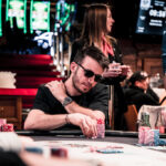 Ein Mann und eine Frau sitzen an einem Pokertisch und spielen eine Partie Poker. Der Mann hält Karten in der Hand, während die Frau mit ernstem Gesichtsausdruck auf ihre eigenen Karten schaut. In