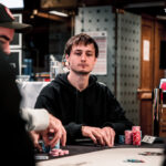 Ein Mann sitzt an einem Pokertisch und hält Karten in der Hand. Er trägt eine dunkle Brille und konzentriert sich auf das Spiel. Im Hintergrund sind weitere Spieler und Chips zu sehen.