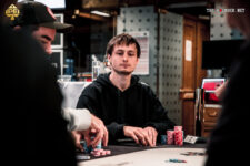 Ein Mann sitzt an einem Pokertisch und hält Karten in der Hand. Er trägt eine dunkle Brille und konzentriert sich auf das Spiel. Im Hintergrund sind weitere Spieler und Chips zu sehen.