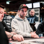 Ein Mann in einem schwarzen Anzug und einer schwarzen Sonnenbrille spielt Poker an einem Tisch in einem eleganten Casino. Er hält Karten in der Hand und schaut konzentriert auf den Tisch. Um